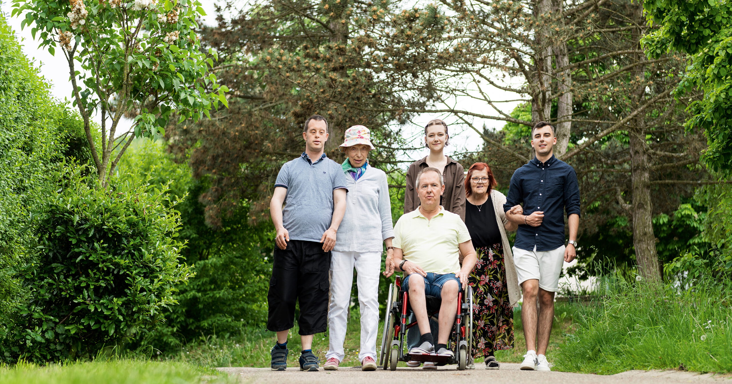 Auf einem grünen Pfad in der Nähe des Sproll-Hauses genießt eine Gruppe von sechs Personen die Natur. Eine Person sitzt im Rollstuhl, sie gehen gemeinsam spazieren und lächeln. Sie verkörpern die freundliche, entspannte Atmosphäre, die die Treffen von Bischof Sproll auszeichnet.