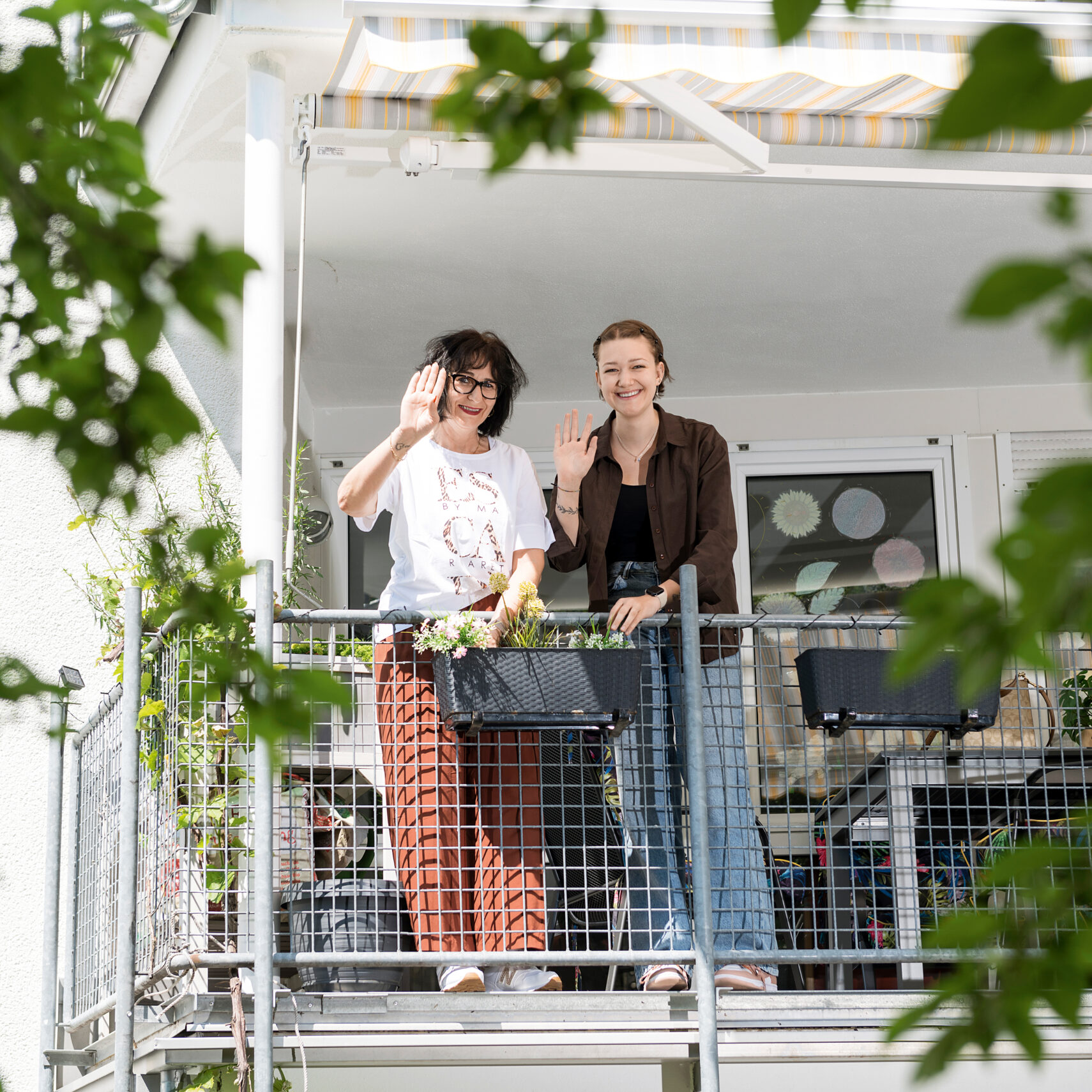 Zwei Menschen stehen auf einem Balkon des Bischof-Sproll-Hauses, lächeln und winken. Sie sind umgeben von Topfpflanzen und grünem Blattwerk. Über ihnen ist eine gestreifte Markise, hinter ihnen befinden sich Glastüren.