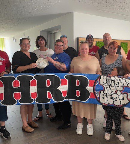 Eine Gruppe von Menschen steht drinnen zusammen, lächelt und hält ein buntes Banner mit dem Text „HRB“. Eine Person hält einen kleinen, in Stoff gewickelten Gegenstand. Die Gruppe wirkt lässig und entspannt, als würde sie eine wohltätige Spende für das Hospiz Barbara feiern.