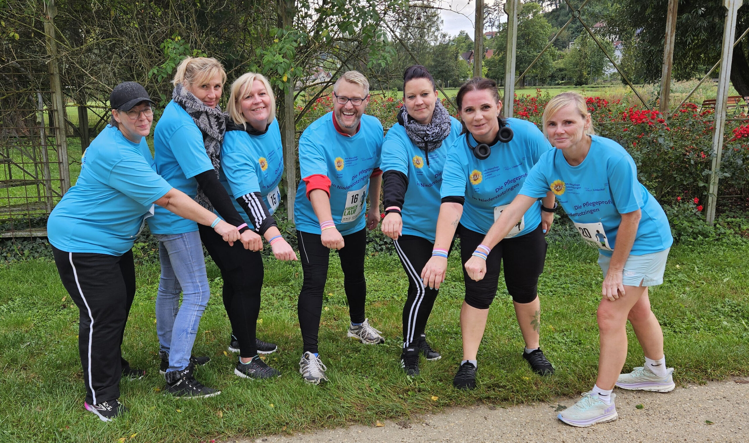 Sieben Personen in passenden blauen Shirts und Startnummern posieren in einer Reihe im Freien, jeder streckt einen Arm nach vorne. Sie feiern ihre Teilnahme am Firmenlauf auf einer Wiese neben einem Weg, mit Bäumen und Büschen im Hintergrund, und präsentieren voller Stolz ihr tolles Ergebnis.