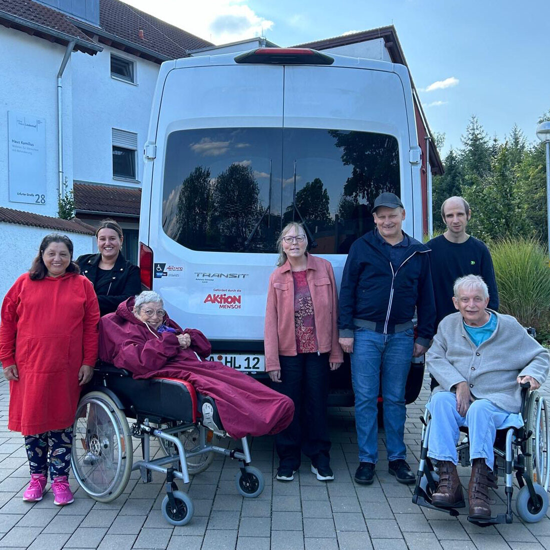 Eine Gruppe von sieben Menschen, darunter zwei im Rollstuhl, steht lächelnd vor einem weißen Lieferwagen mit der Aufschrift „Neuer Haus Kamillus“. Die Szene ist im Freien mit einem Gebäude und Bäumen im Hintergrund.