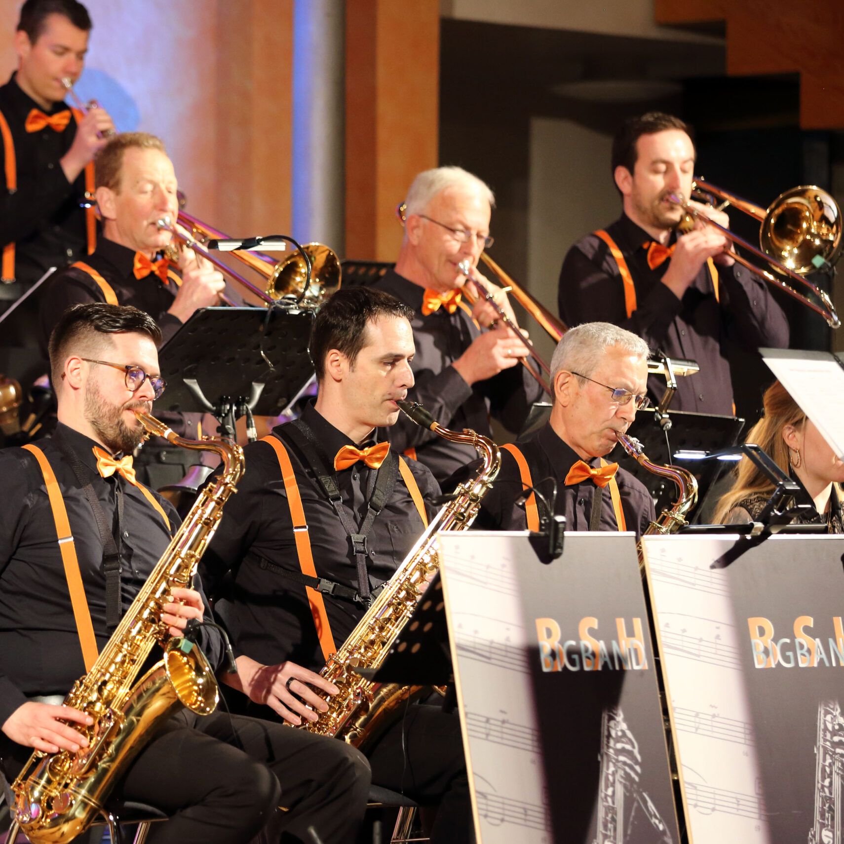 Bei einem Benefizkonzert steht die BSH Big Band mit sechs Musikern, überwiegend Männern, auf der Bühne, die Saxophone und Trompeten spielen. Sie tragen schwarze Hemden mit orangefarbenen Hosenträgern und Fliegen und treten vor Notenständern auf, auf denen vor einem schwach beleuchteten Hintergrund „RGBand“ steht.