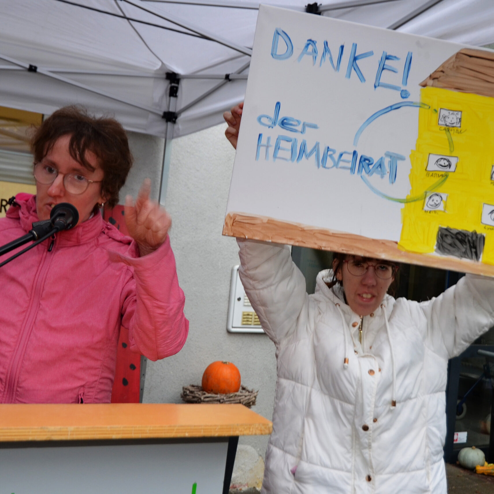 Bei der Open-Air-Veranstaltung zum 25-jährigen Jubiläum spricht eine Person in einer rosa Jacke auf einem Podium, während eine andere in einer weißen Jacke stolz ein Schild mit handgeschriebenem Text und Skizzen hochhält. Hinter ihnen wird die festliche Atmosphäre durch einen kleinen Kürbis auf dem Tisch unterstrichen.