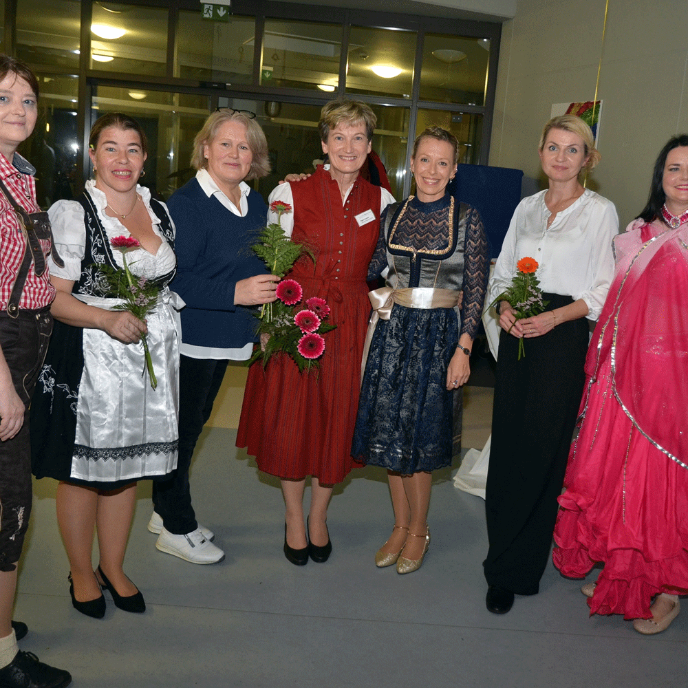 Sieben Frauen stehen lächelnd und mit Blumen in den Händen in einem Flur des Ehrenamtsfestes. Sie tragen verschiedene traditionelle und formelle Gewänder vor einer Fensterkulisse und sanfter Innenbeleuchtung.