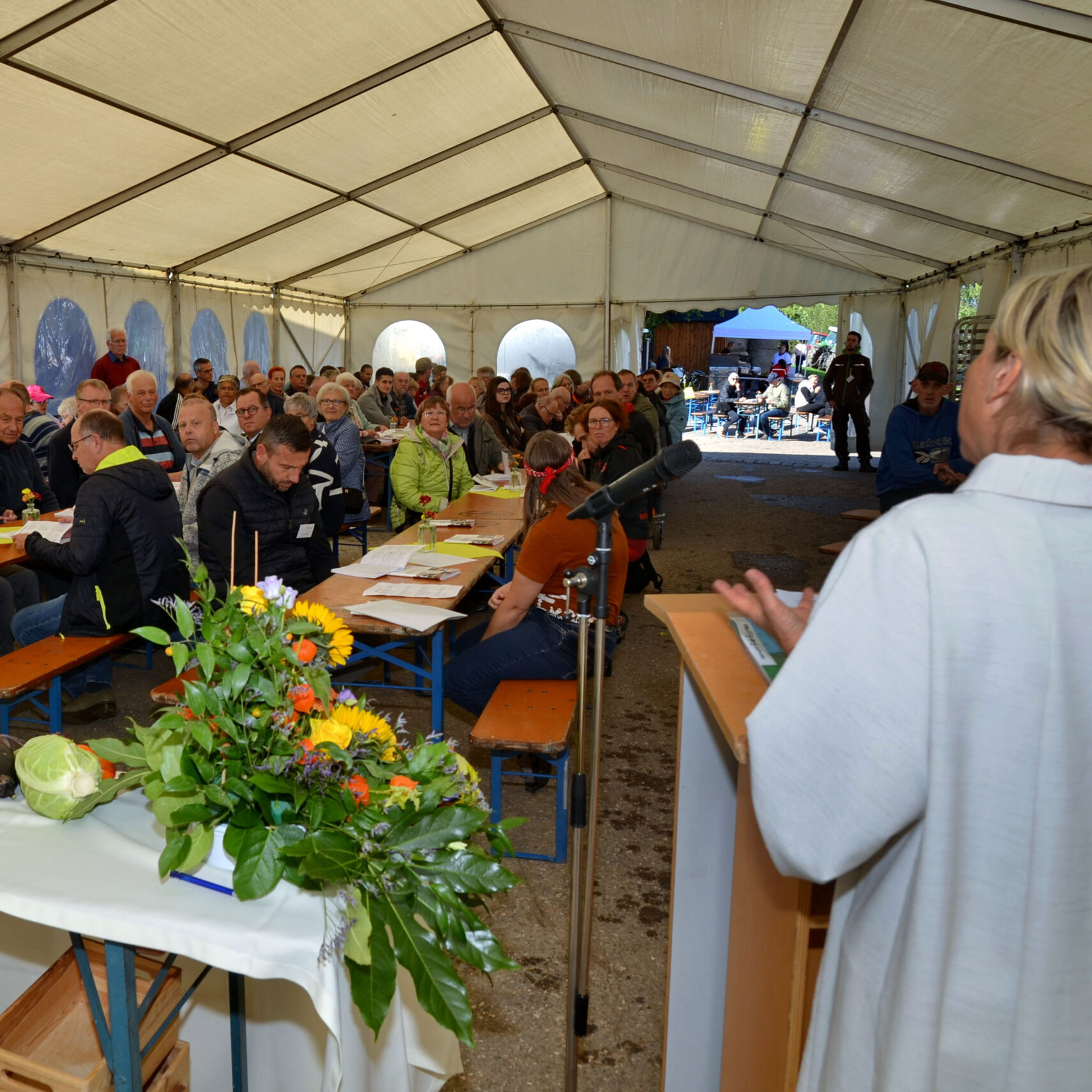 Beim Haldenhoffest hält eine Person auf einem Podium in einem großen Zelt eine Rede. Ein Publikum, das an langen Tischen sitzt, hört aufmerksam zu. Ein farbenfrohes Blumenarrangement schmückt einen Tisch im Vordergrund und trägt zur festlichen Atmosphäre dieses geschätzten Ereignisses bei.