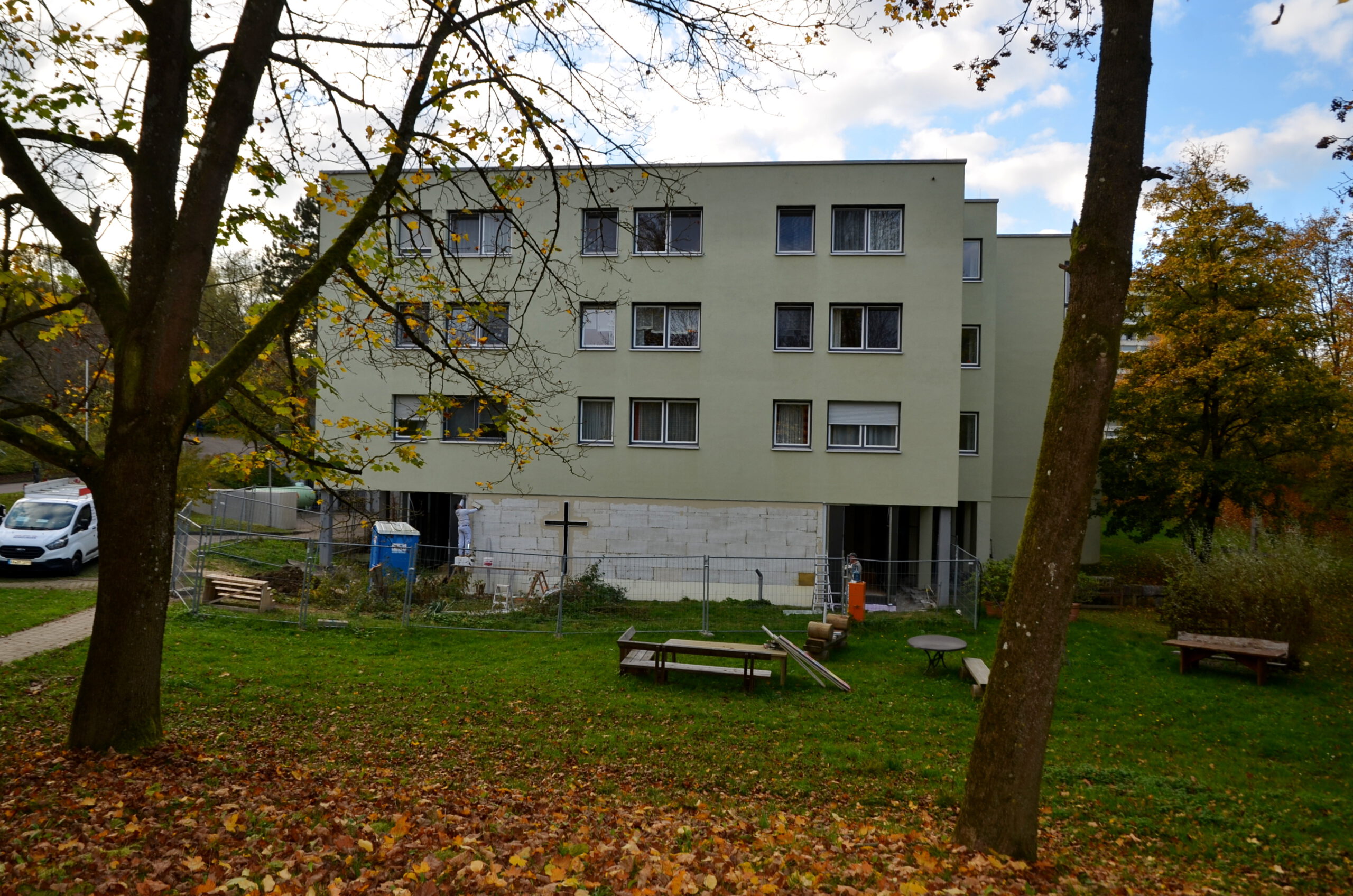 Ein hellgrünes, dreistöckiges Mehrfamilienhaus steht zwischen herbstlich gefärbten Bäumen. Davor sind eine Rasenfläche mit Bänken und ein kleines Baufortschrittsgelände zu sehen, links ein teilweise geparkter weißer Lieferwagen.