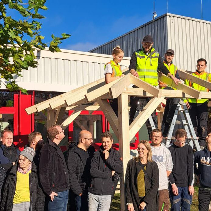 Unter sonnigem Himmel steht eine Gruppe der Christophorus-Werkstatt in Freizeitkleidung und Warnwesten auf Leitern und baut eine hölzerne Dachkonstruktion für den Neuen Pavillon. Unten schaut eine Menschenmenge gespannt zu. Ein Baum und ein Gebäude rahmen diese lebhafte Szene ein.