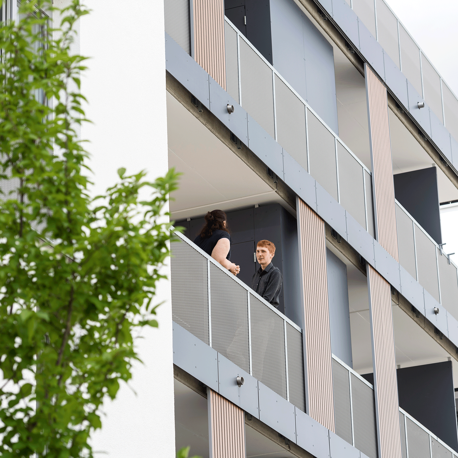 Zwei Menschen stehen und unterhalten sich auf einem Balkon eines modernen Hochhauses in Heidenheim. Die Struktur besteht aus vertikalen und horizontalen Paneelen, im Vordergrund ist teilweise ein Baum zu sehen, der an eine architektonische Beratungsstelle erinnert.