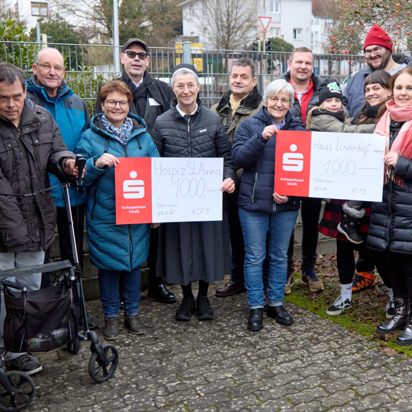 Eine Gruppe von Menschen hält im Freien Banner mit Spendenbeträgen für das „Hospiz St. Anna“ und das „Haus Lindenhof“ hoch, beide zeigen „1000“. Gemeinsam posieren sie lächelnd am Adventsweg in Röhlinger, inmitten einer Kulisse aus Bäumen und einem Metallzaun.