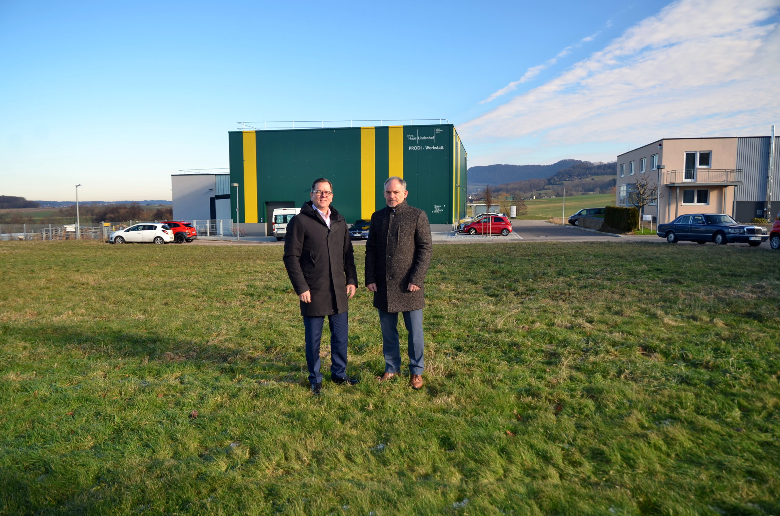 Zwei Männer stehen auf einer Wiese vor einem großen grün-gelben Industriegebäude. Unter dem klaren blauen Himmel bilden die entfernten Hügel eine ruhige Kulisse. Auf der rechten Seite ergänzen Autos und ein kleines Gebäude diese automatisch gestaltete Szene.