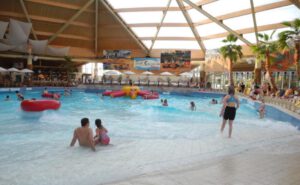 Der Indoor-Wasserpark erinnert an Palm Beach und verfügt über ein Wellenbad, in dem die Menschen sich im Wasser vergnügen können. Umgeben von Palmen unter einer Holzbalkendecke bietet Nürnbergs Wasseroase aufblasbare Schläuche und eine Spielstruktur, während im Hintergrund Liegestühle zum Entspannen einladen.