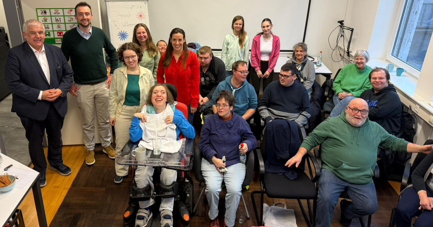 Eine bunt gemischte Gruppe von Menschen, einige davon im Rollstuhl, posiert gemeinsam in einem hellen Raum. Im Hintergrund ist eine Tafel mit bunten Zeichnungen zu sehen. Sie lächeln und scheinen sich zu einem Workshop zum Thema „einfach erklärt Politik“ versammelt zu haben.