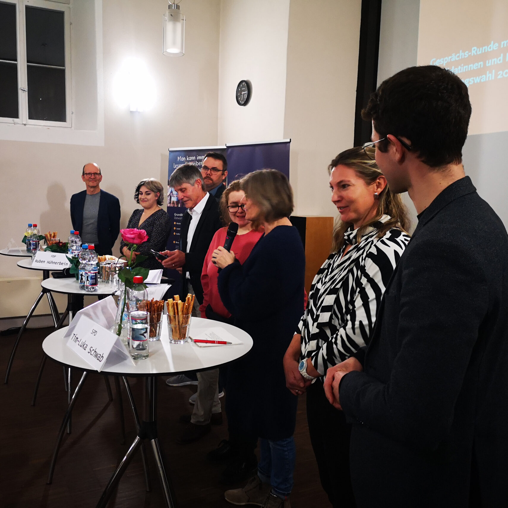 Eine Gruppe von Menschen steht in einer Reihe in einem Raum mit hohen Fenstern. Eine Frau spricht in ein Mikrofon und erklärt einfach Politik. Auf den Tischen vor ihnen stehen verschiedene Dinge wie Getränke und Snacks. Die Umgebung scheint eine formelle oder professionelle Veranstaltung zu sein.