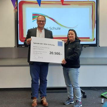 Zwei Personen stehen in einem Klassenzimmer und halten einen großzügigen Scheck über 20.500 Euro der IKEA-Stiftung in den Händen. Im Hintergrund ist auf einem Bildschirm der Schriftzug „Markus Schule“ zu sehen. Der Raum ist mit farbenfrohen Dekorationen geschmückt, die die großzügige Spende feiern.