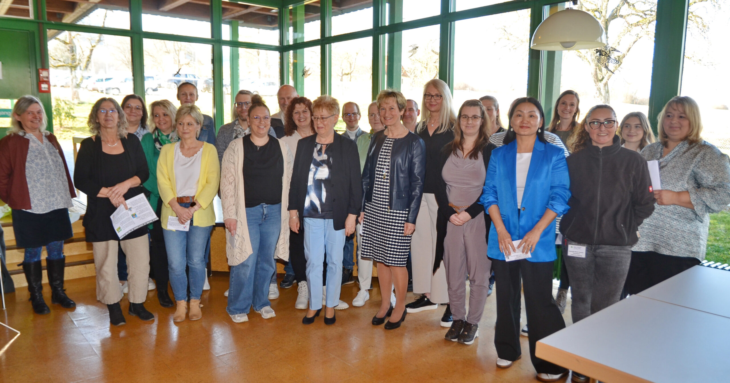 Eine bunt gemischte Gruppe von Menschen steht in einem hellen Raum mit großen Fenstern zusammen. Sie sind leger gekleidet, lächeln und halten Papiere in den Händen, was auf eine HLS-Versammlung schließen lässt. Die üppigen grünen Bäume im Hintergrund verstärken die warme Atmosphäre dieser Veranstaltung rund um einen Leitungswechsel.